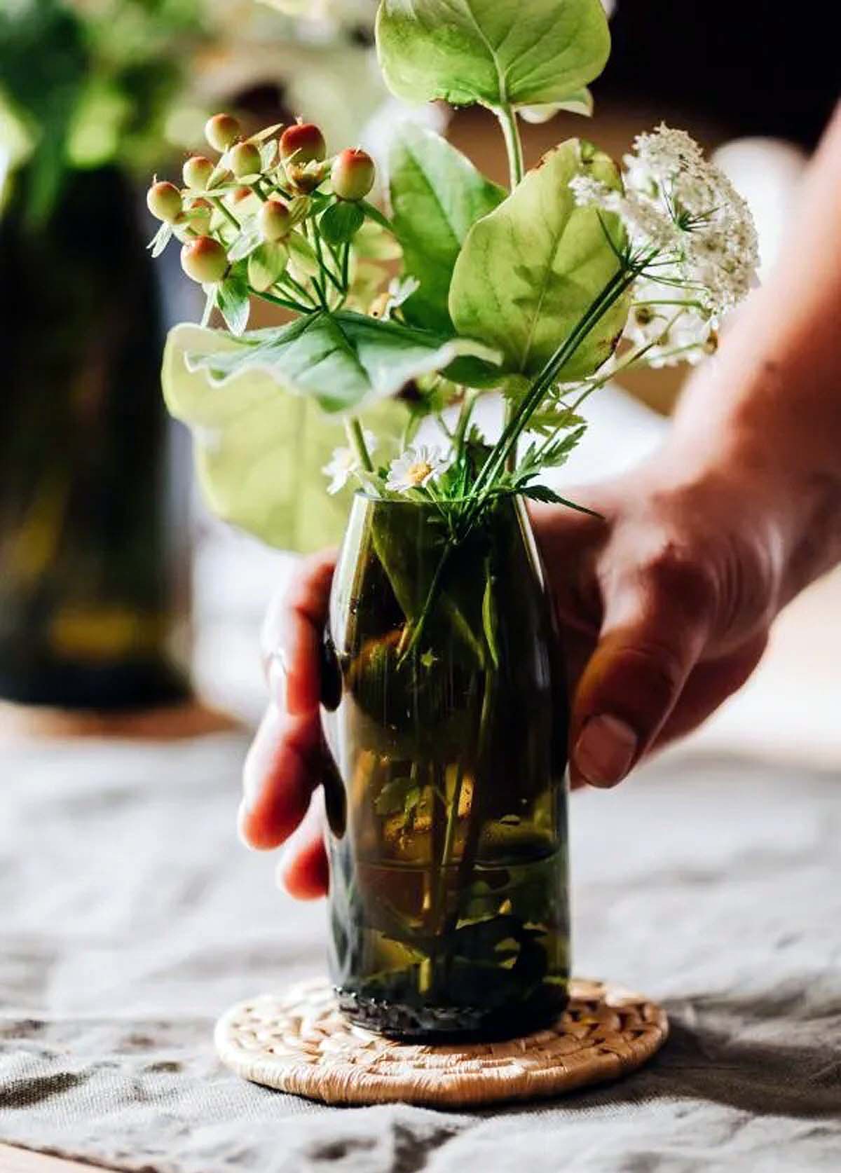 olive-green-piccolo-glass-vase hand placing small vase filled with flowers onto a coaster on a covered table