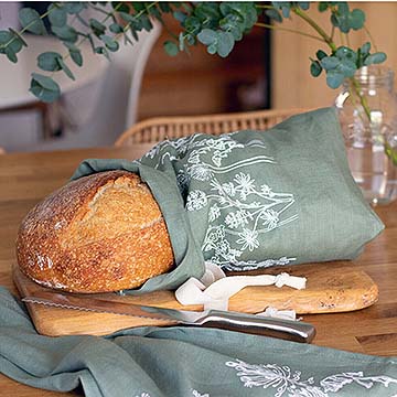 sage-green-bread-bag loaf of bread peeping out from bread bag on bread board with bread knife on work surface